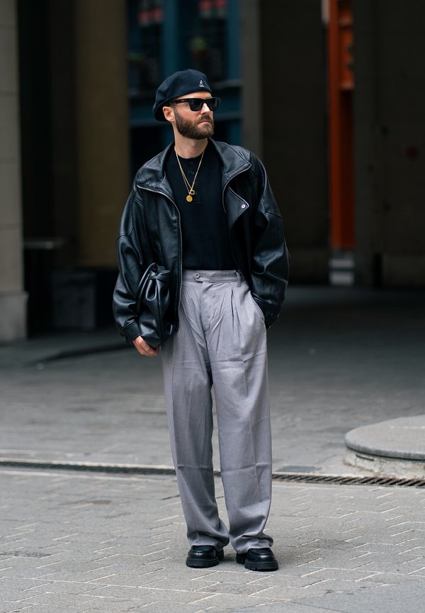 Un hombre barbudo con gafas de sol, una chaqueta de cuero negra y una boina, y unos pantalones grises holgados está de pie en una acera de la ciudad.