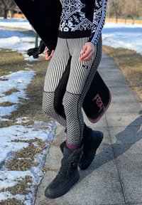 Personne en leggings à motifs et bottes d'hiver noires portant un snowboard noir sur un chemin extérieur enneigé.