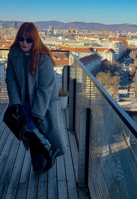 Kobieta o długich rudych włosach, w ciemnych okularach przeciwsłonecznych, szarym płaszczu, rękawiczkach i czarnych butach stoi na miejskim balkonie z widokiem na góry w świetle dziennym.
