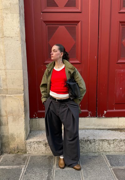 Persona con cabello oscuro recogido hacia atrás, de pie junto a una puerta roja, lleva una chaqueta verde, suéter rojo, pantalones negros holgados, zapatos marrones y sostiene una bolsa negra.