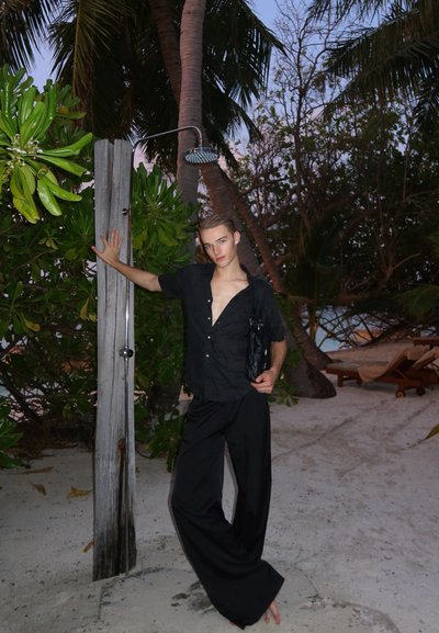 Camisa negra de manga corta con cuello abierto y pantalones anchos negros, sosteniendo una bolsa negra. Rodeada de vegetación cerca de una ducha y arena.