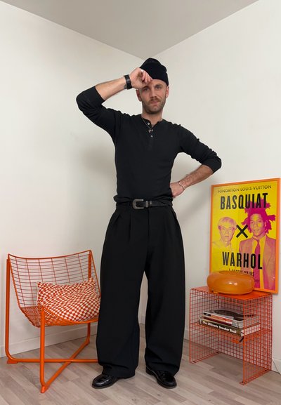 Hombre con atuendo negro y gorro de lana de pie en una habitación minimalista con una silla de alambre naranja, cojín a cuadros, mesa auxiliar naranja y un póster de Basquiat Warhol.