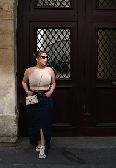 Top halter de punto beige, falda maxi de mezclilla oscura con abertura frontal, zapatillas blancas y un pequeño bolso de crema. De pie junto a una puerta de madera oscura con un patrón de rejilla.