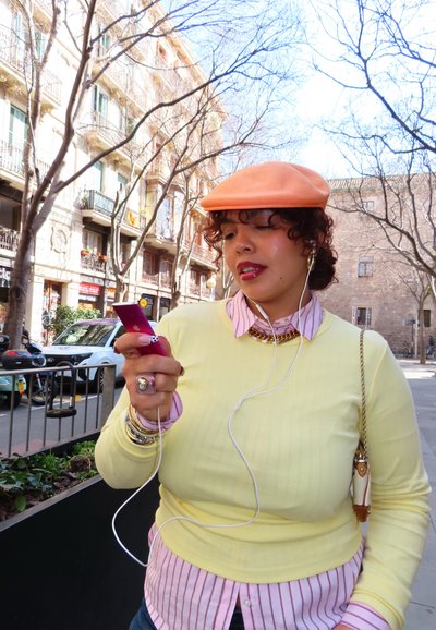 Mujer con boina melocotón y suéter amarillo usando un teléfono inteligente con auriculares en una calle de la ciudad, con árboles sin hojas y edificios de fondo.