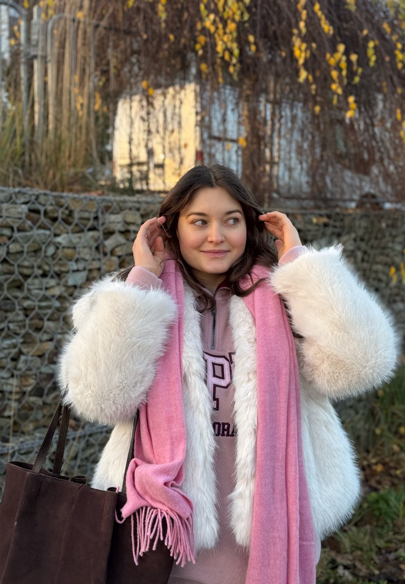 Manteau en fausse fourrure blanche avec écharpe rose, sweat à capuche gris et sac fourre-tout marron. Matériaux texturés et couleurs chaudes. Cadre extérieur avec un arrière-plan de feuillage.