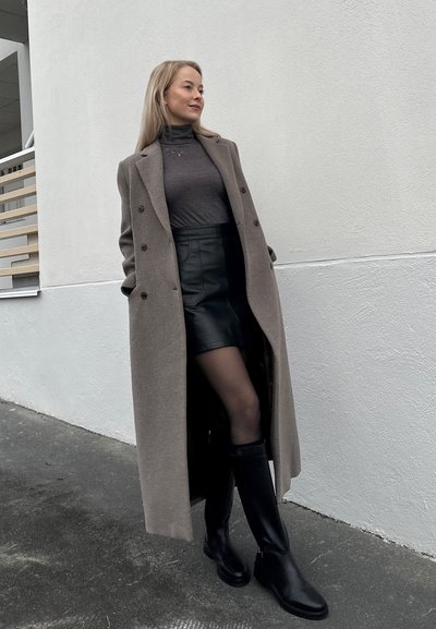Abrigo largo, de doble botonadura, de lana gris sobre una blusa ajustada oscura y una minifalda de cuero. Botas altas negras hasta la rodilla completan el atuendo.