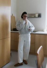 Pull gris flou superposé sur un haut crème ; pantalon blanc et chaussures marron ; textures douces ; arrière-plan de cuisine minimaliste.