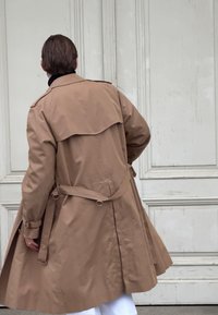 Manteau trench beige avec un design croisé à double boutonnage, grand rabat au dos, taille ceinturée et revers roulés, vu de dos contre un mur texturé.