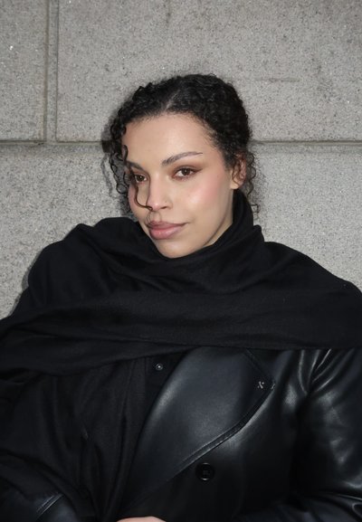 Joven con cabello rizado, envuelta en una bufanda negra y chaqueta de cuero, de pie contra una pared de piedra gris, mirando ligeramente hacia un lado.