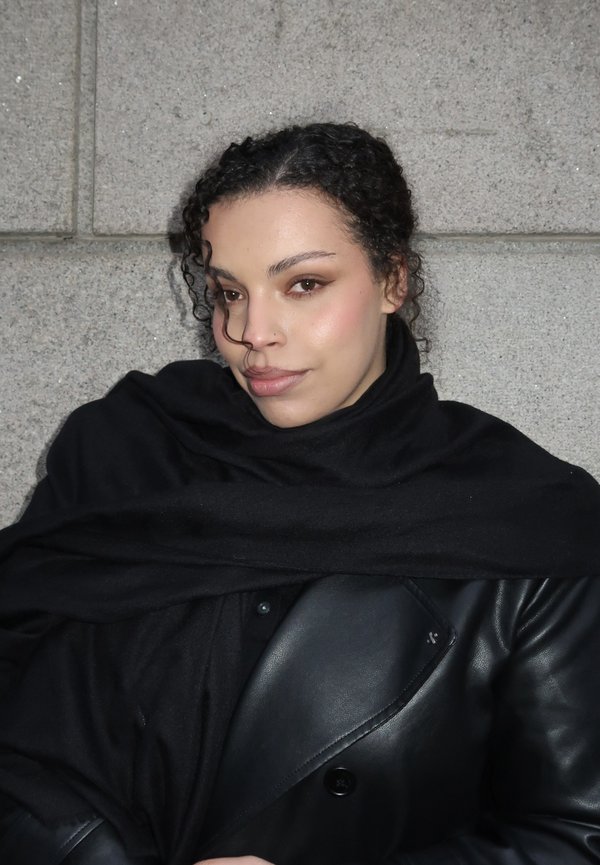 Ung kvinne med krøllete hår pakket inn i et svart skjerf og skinnjakke, stående foran en grå steinvegg, ser litt til siden.