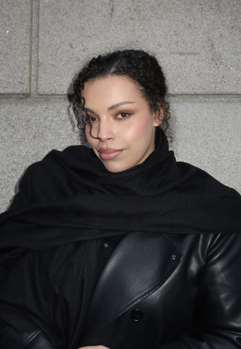 Jeune femme aux cheveux bouclés enveloppée dans une écharpe noire et une veste en cuir, debout contre un mur en pierre grise, regardant légèrement sur le côté.