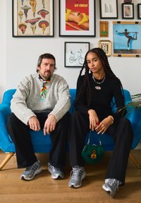 Un homme et une femme assis sur un canapé bleu, tous deux portant des baskets grises ; le mur derrière eux est décoré avec des œuvres d'art éclectiques encadrées et des affiches.