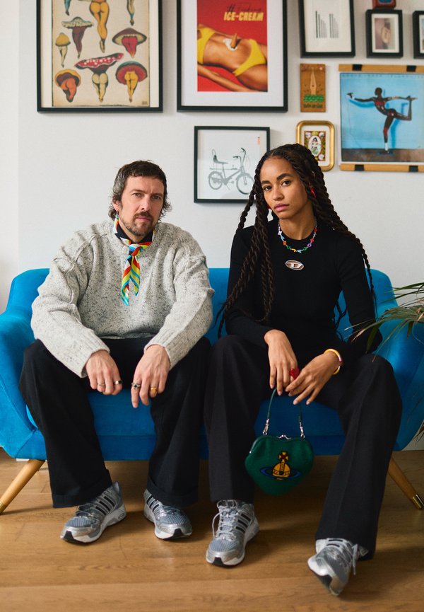 Uomo e donna seduti su un divano blu, entrambi indossano sneaker grigie; la parete dietro di loro è decorata con opere d'arte e poster eclettici incorniciati.