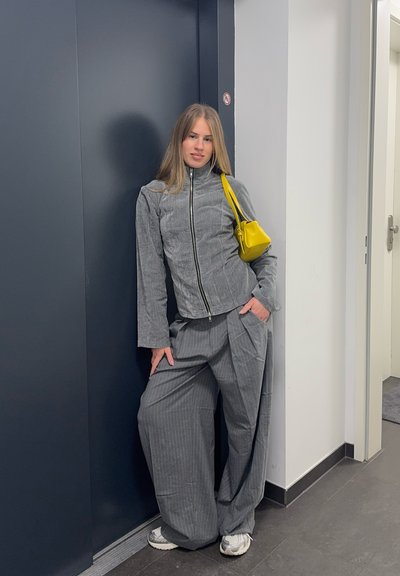 Mujer joven con chaqueta gris con cremallera y pantalones anchos gris, apoyada contra una pared azul oscuro, llevando un pequeño bolso amarillo.