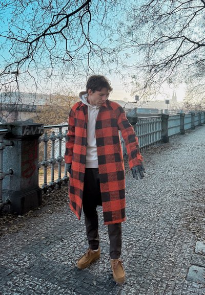 Abrigo de lana a cuadros en rojo y negro con botones, combinado con una sudadera blanca y pantalones negros. El conjunto incluye zapatos marrones y guantes negros.