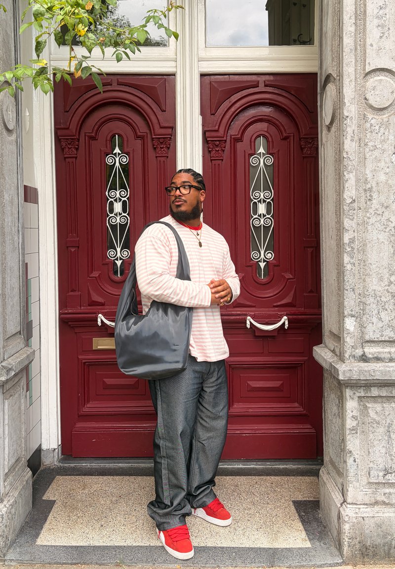 Homme avec des lunettes, cheveux tressés, chemise rose à rayures, pantalon gris, chaussures rouges et grand sac noir se tient devant des portes doubles rouges ornées.