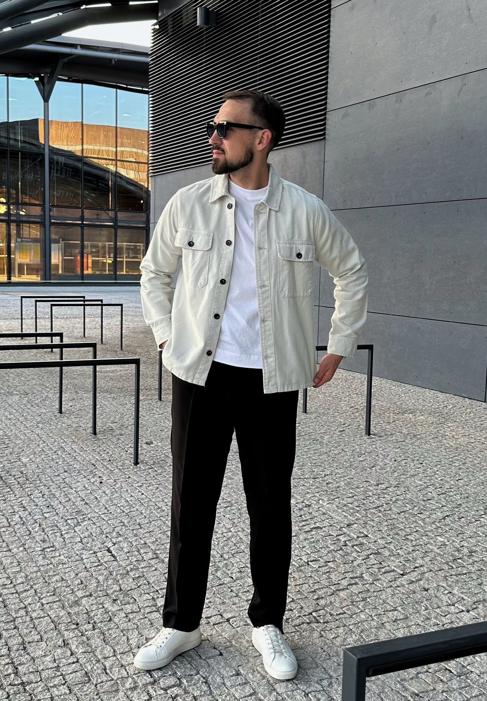 Gaišas žakete ar melniem pogām, baltā T-kreklā, melnās bikses un baltas kedas. Āra vide ar mūsdienīgu arhitektūru fona.