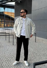 Gaišas žakete ar melniem pogām, baltā T-kreklā, melnās bikses un baltas kedas. Āra vide ar mūsdienīgu arhitektūru fona.