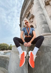 Zapatillas de correr naranjas con suelas y cordones blancos, leggings negros y una camiseta corta gris. El fondo presenta arquitectura clásica.