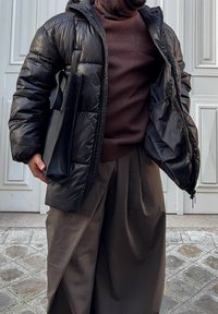 Veste matelassée noire avec finition brillante, pull marron en dessous et pantalon marron ample. Tient un sac noir. Fond en pavé de pierre.