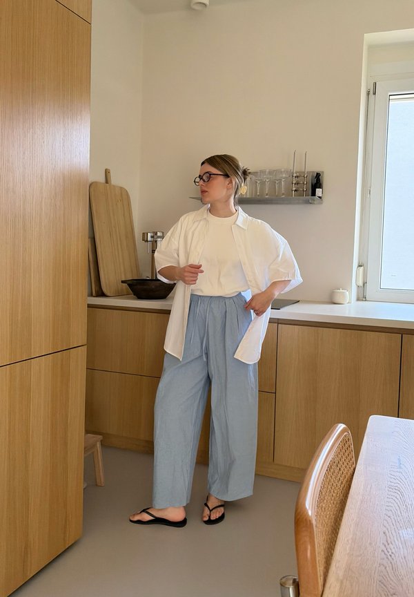 Donna con occhiali che indossa una camicia bianca e pantaloni azzurri in piedi in una cucina minimalista con mobili in legno e una finestra.
