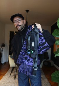 Mann med briller og caps holder en svart veske og et lilla skjerf med mønster inne i en stue.