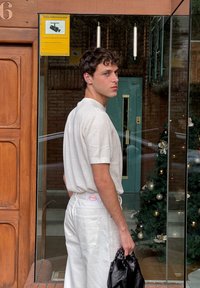 Witte textuur korte mouwen shirt, witte broek met hoge taille en een zichtbaar logo aan de achterkant, en een zwarte handtas, tegen een glazen etalage.