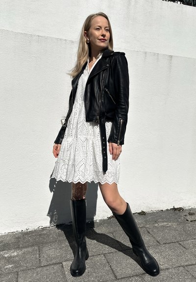 Chaqueta de cuero negro tipo motero superpuesta sobre un vestido blanco de encaje con dobladillo de ondas. Botas negras hasta la rodilla completan el conjunto.