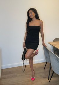 Black strapless dress, fitted silhouette, with a smooth texture. Pink stiletto heels with bead accents. Holding a black handbag.