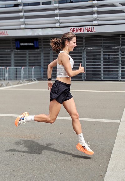 Atleta con una camiseta sin mangas clara y pantalones cortos negros corriendo sobre el pavimento. Lleva zapatillas New Balance con acentos naranjas y calcetines blancos.