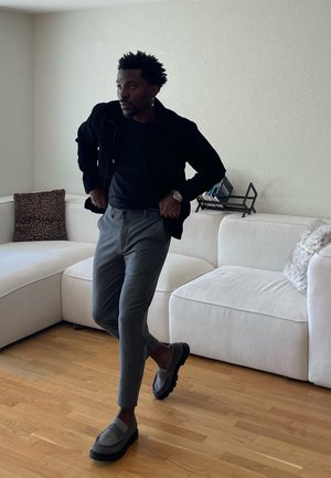 Hombre con chaqueta negra y pantalones grises caminando en el interior cerca de un sofá seccional blanco con cojines, suelo de madera y libros en un estante.