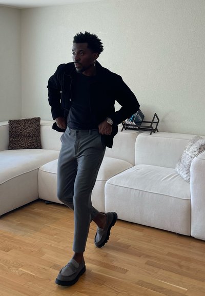 Hombre con chaqueta negra y pantalones grises caminando en el interior cerca de un sofá seccional blanco con cojines, suelo de madera y libros en un estante.