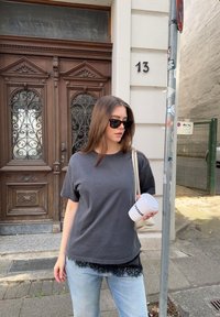 Tricou gri din bumbac cu dantelă, purtat cu blugi deschiși la culoare. Ochelari de soare și o cană albă de cafea cu textul "venue". Ușă decorativă în fundal.