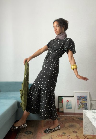 Mujer con vestido negro de lunares equilibrándose sobre un pie cerca de un sofá azul, sosteniendo una bolsa verde con una obra de arte enmarcada apoyada contra la pared.