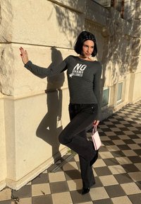 Femme aux cheveux noirs coupés au carré portant un t-shirt rayé avec l'inscription « NO DRAMA ALLOWED », un jean noir et un bandeau, tenant un sac à main rose, appuyée contre un mur en pierre.