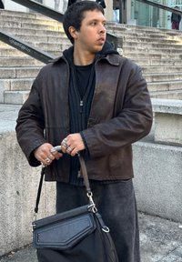 Brown leather jacket, black zip-up sweater, and dark trousers. Carrying a black bag with a textured flap and silver hardware. Adorned with various silver rings.