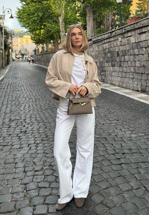 Chaqueta corta beige sobre una camisa blanca, combinada con pantalones blancos acampanados. Sostiene un pequeño bolso gris, de pie en una calle de adoquines.