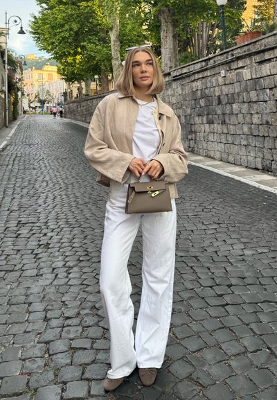 Chaqueta corta beige sobre una camisa blanca, combinada con pantalones blancos acampanados. Sostiene un pequeño bolso gris, de pie en una calle de adoquines.