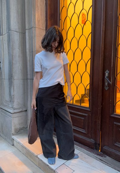 Camisa blanca de manga corta, falda negra sobre jeans oscuros de pierna ancha, sosteniendo un bolso marrón, con una ventana arqueada decorativa de fondo.