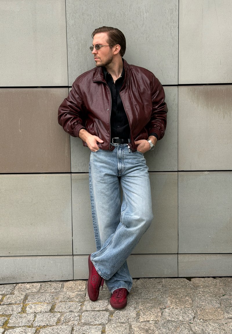 Homme portant une veste en cuir bordeaux, une chemise noire, un jean bleu clair et des baskets rouges, appuyé contre un mur carrelé avec les mains dans les poches.