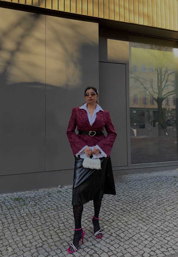 Woman wearing maroon textured blazer, white shirt, black leather skirt, patterned tights, pink heels, sunglasses, and holding a small white handbag.