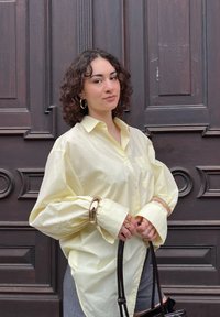 Camisa oversized botão-amarelo claro com mangas longas, combinada com calças cinzentas. Acessórios dourados notáveis e uma bolsa preta.