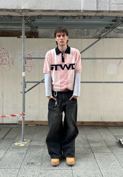 Polo a rayas rosa y blanca con cuello negro y logotipo, superpuesto sobre una camisa de manga larga gris; combinado con jeans oscuros de corte amplio y zapatos marrones.