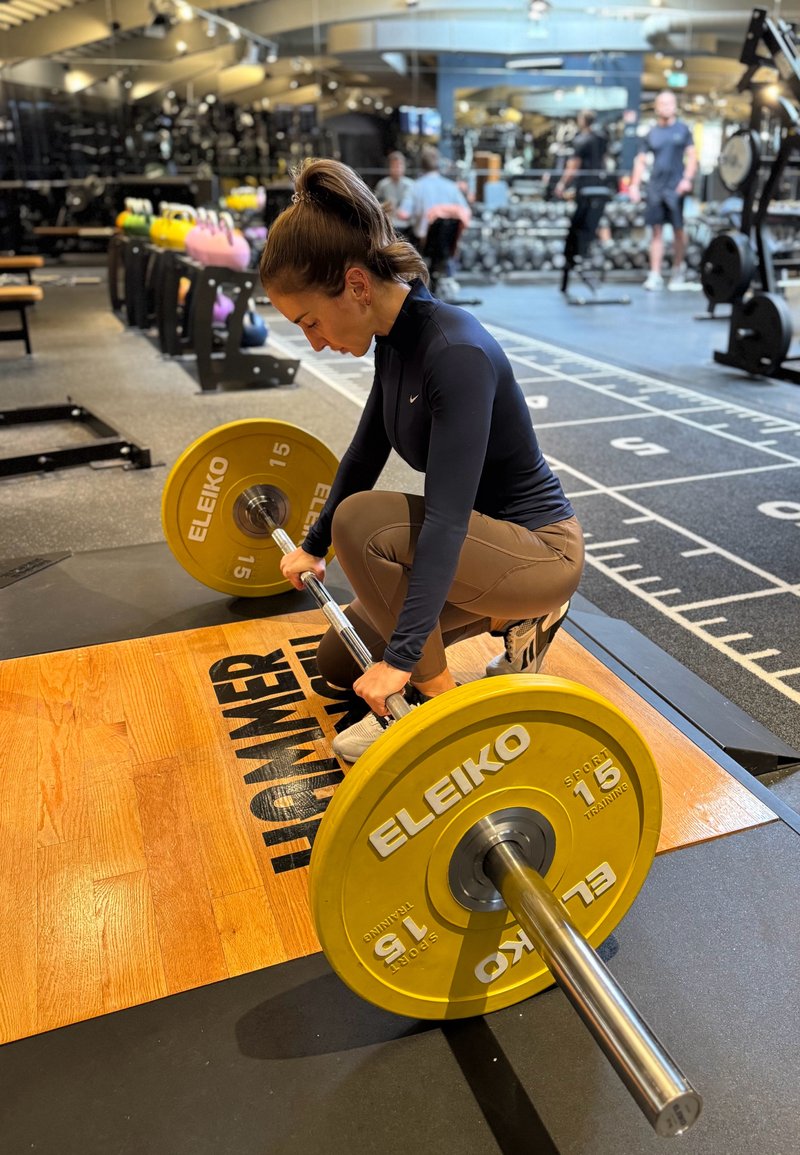 Barra con discos de pesas ELEIKO amarillos (15 kg) sobre una plataforma de madera. Una persona se agacha, sujetando la barra con ambas manos. Entorno de gimnasio.