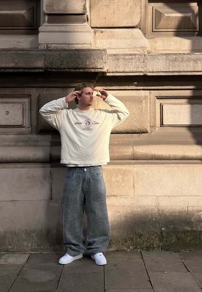 Joven con sudadera blanca y vaqueros anchos ajustándose el cabello, de pie frente a una pared de piedra texturizada iluminada por el sol sobre un suelo pavimentado.