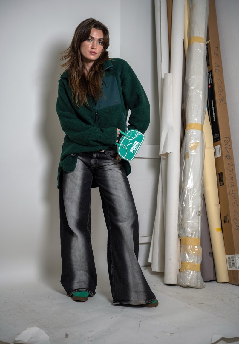 Giovane donna con un ampio pile verde scuro e jeans larghi neri sbiaditi in piedi accanto a tele arrotolate, che tiene una piccola borsa verde Puma.