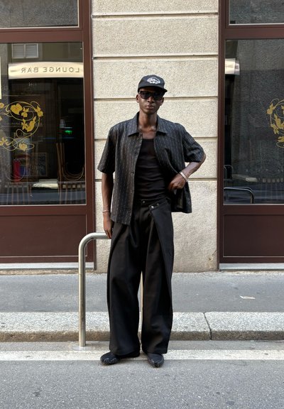 Camisa negra de manga corta con rayas blancas horizontales, camiseta negra sin mangas, pantalones anchos negros y zapatos negros texturizados; entorno urbano.