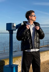 Homme en doudoune noire et chemise rayée portant des lunettes de soleil, debout près d'un télescope bleu sur une promenade au bord de l'eau au coucher du soleil.