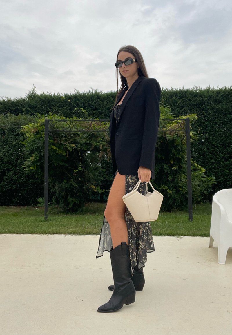 Blazer nero sopra un vestito floreale con uno spacco alto, abbinato a stivali neri al ginocchio e a una piccola borsa crema. Ambientazione all'aperto con vegetazione.