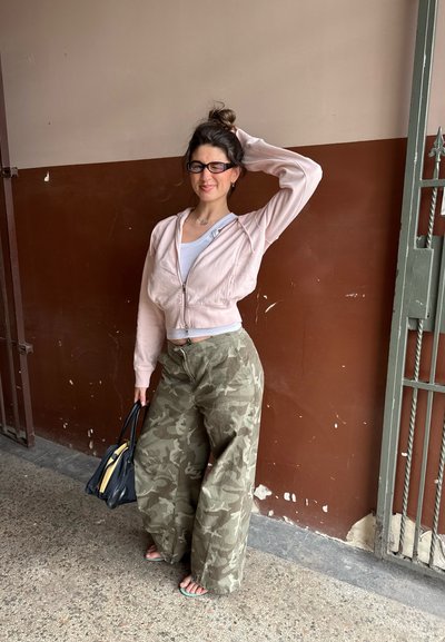 Mujer con gafas lleva sudadera con cremallera rosa claro, camiseta blanca, pantalones anchos de camuflaje, sostiene bolso negro, de pie junto a una pared marrón y beige.